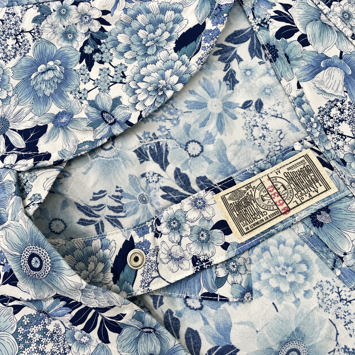 Close-up of a blue and white floral print fabric garment, showing a buttoned pocket with a beige brand label that reads 'KIKO KIKO'.