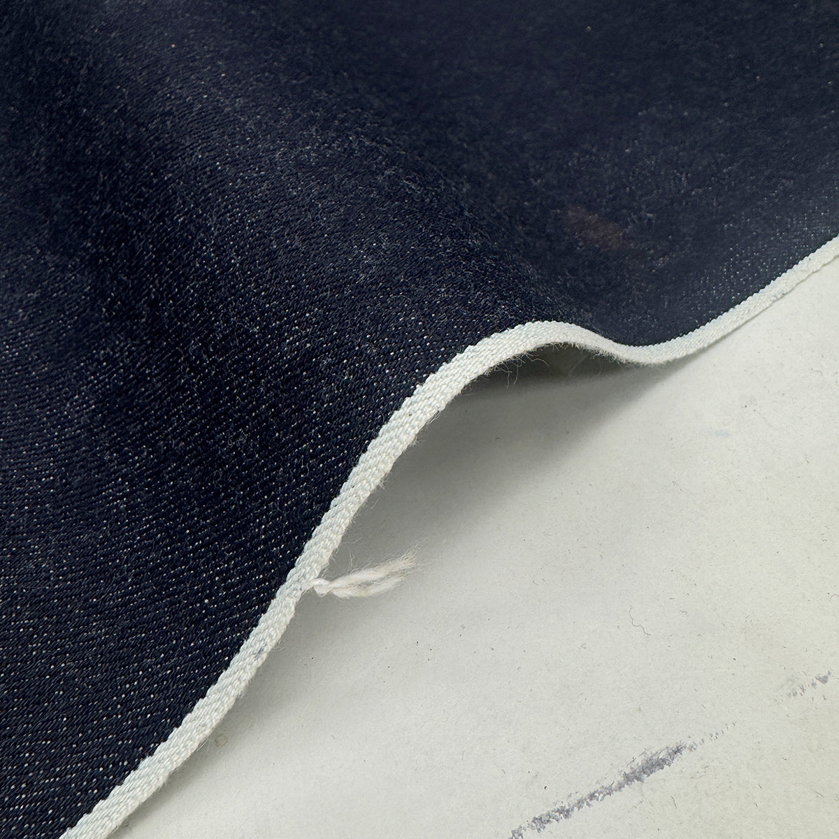 A close-up of dark indigo denim fabric with a white twisted edge, draped over a light gray surface.