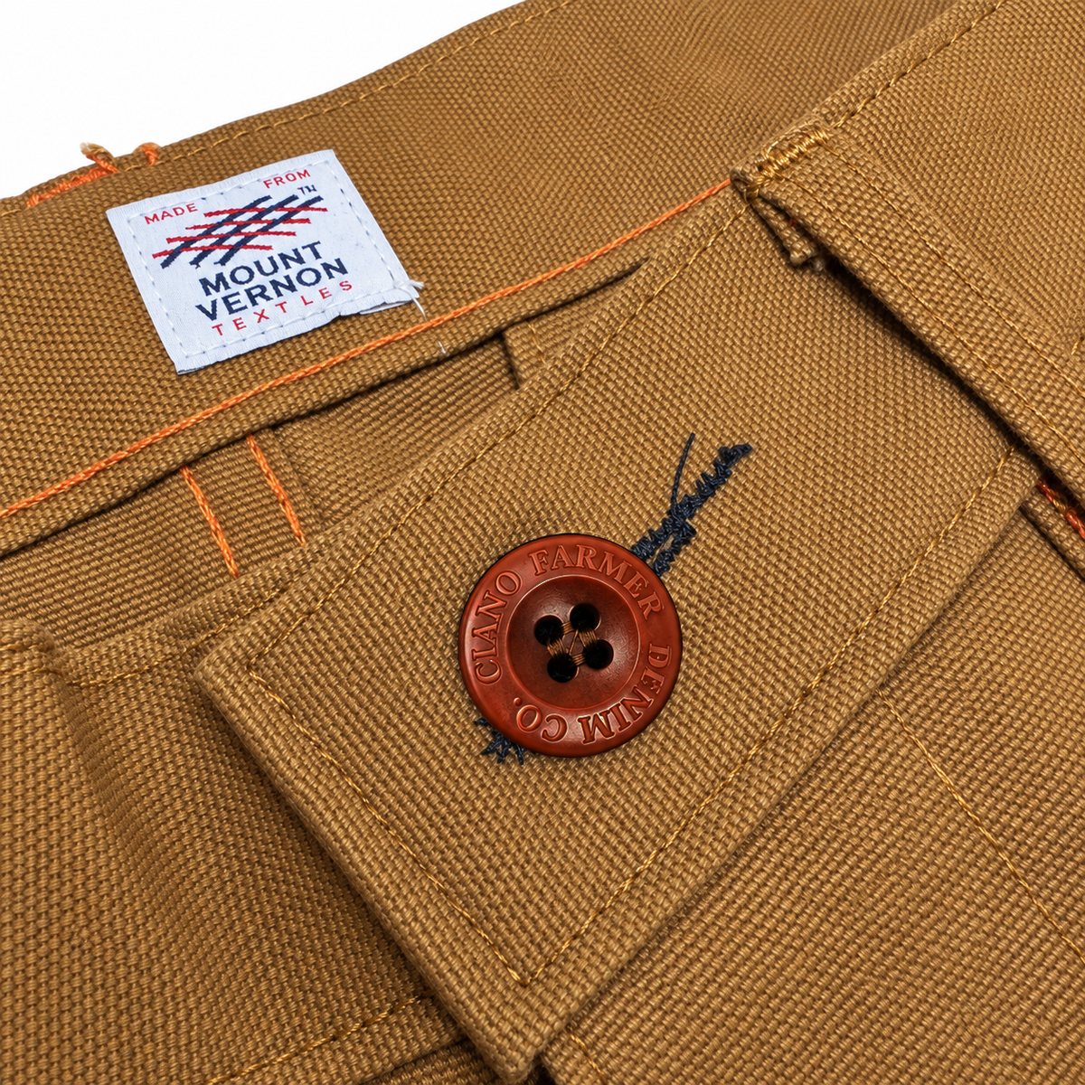Close-up of tan canvas work pants showing a brown buttoned pocket flap, orange contrast stitching, and a white Mount Vernon Tailor label.