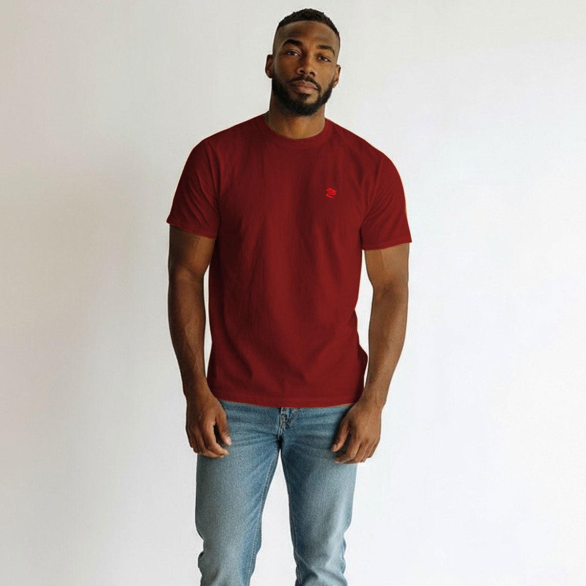 Man wearing a red t-shirt with a small logo and light blue jeans against a plain background