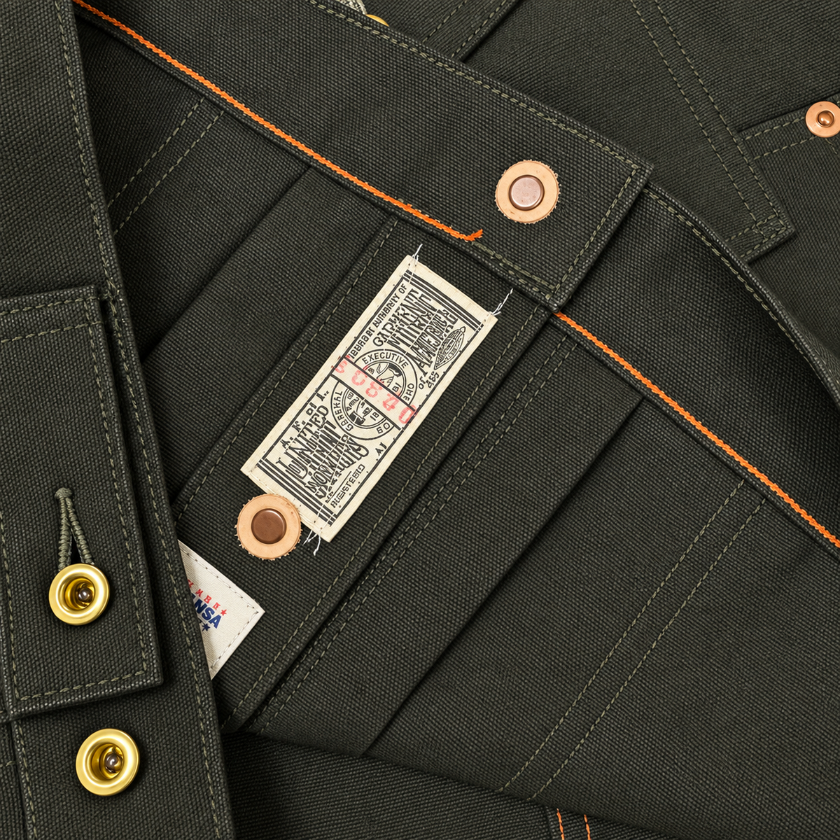 Close-up of dark olive green canvas workwear pants showing the interior waistband construction, orange contrast stitching, brass button details, and a sewn-in paper brand label.